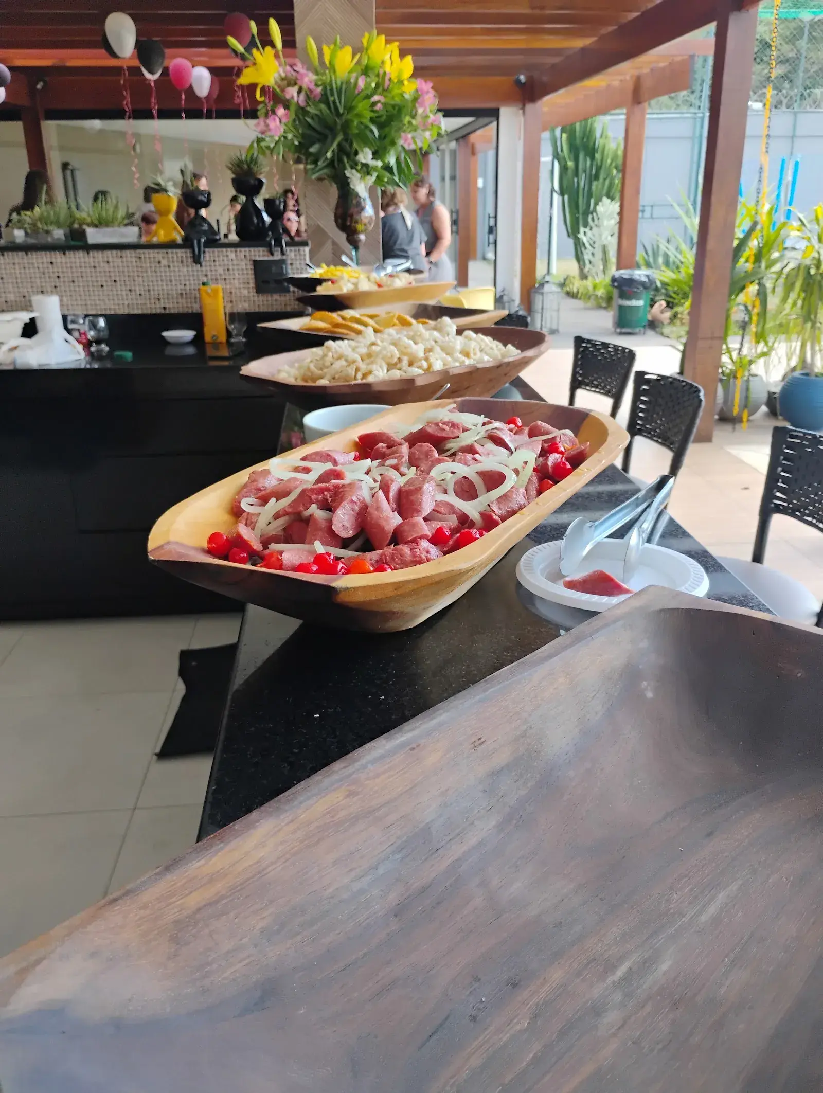 Gamela de linguiça com cebola e pimenta biquinho no buffet, com torresmo e mandioca ao fundo